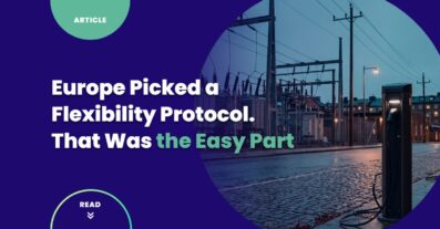 Public EV charge point on a wet cobbled street at dusk in a northern European city, with a medium-voltage distribution substation and overhead lines behind it.