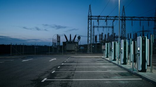 EV charging plaza at dusk with an electrical distribution substation and transmission towers in the background